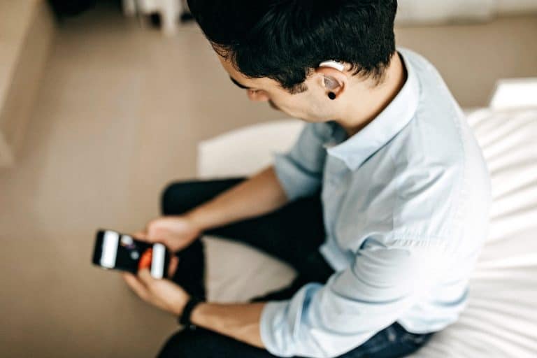 Younger man wearing a hearing aid while looking at his phone and sitting on the edge of a bed