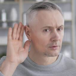 Man in grey shirt in bathroom holding hand up to ear to hear better.