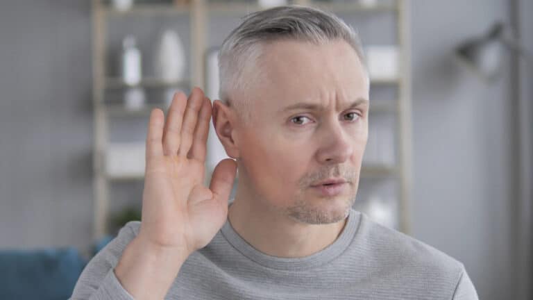 Man having trouble hearing in bathroom. Man in grey shirt in bathroom holding hand up to ear to hear better.