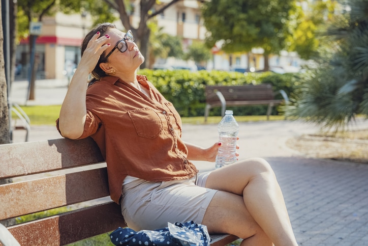 Displeased woman eyes closed sitting on a park bench holding a water bottle. Suffering from heat stroke and headache Displeased woman eyes closed sitting on a park bench holding a water bottle.